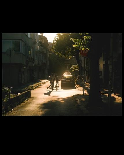 People walking through a sunny spot in a dense urban area. High contrast. Sun is reflecting from the roofs of parked cars. The sunrays make the leaves of invasive plants light up in green. Everything is bloomy.