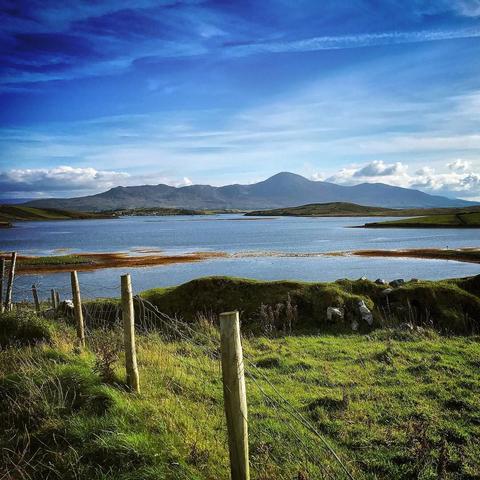 A scenic landscape featuring a calm lake surrounded by lush green hills and a backdrop of mountains under a blue sky with scattered clouds. Wooden fence posts are visible in the foreground.