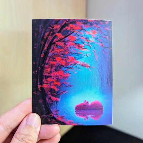 Photo of a left hand holding a rectangle sticker. The sticker is a matte painting of a capybara resting under fall leaves.
