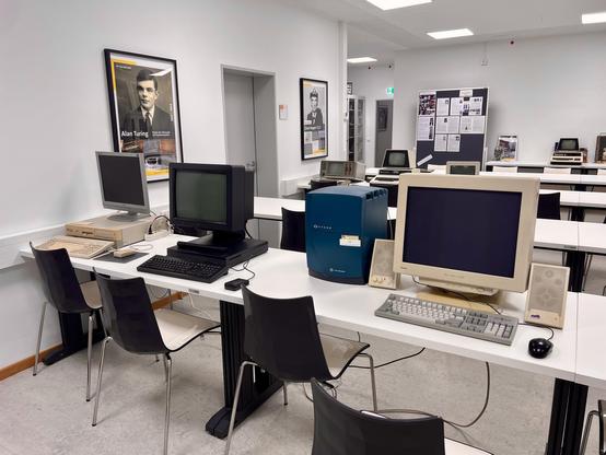 A modern classroom filled with retro computers and technology, with a framed portrait of Alan Turing on the wall. Various computer setups are present on white tables, featuring different styles of monitors and keyboards, alongside a bulletin board displaying historical information.