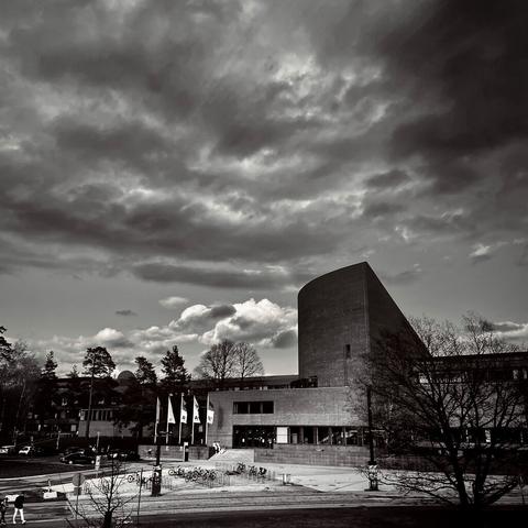 The Undergraduate Centre of Aalto University, surrounded by flags and bicycles, under a dramatic cloudy sky in black and white.