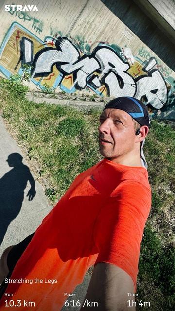 Me taking a selfie while stretching after a run. They are wearing an orange shirt and a black headband. A colorful graffiti wall is visible in the background, and the display shows running statistics: distance of 10.3 km, pace 6:16/km, total time 1h 4m.