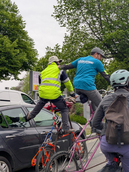 Dans le cortège, deux cyclistes en tallbike à l’arrêt. Le premier s’appuie de son pied sur une voiture en stationnement, le second, qui porte un t-shirt bleu floqué Vélorution, pose sa main sur l’épaule du premier.