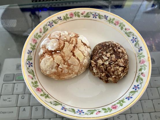 A small plate holds two round treats: one light-colored, powdered, and slightly cracked, and the other dark, round, and textured with oats. The plate features a floral pattern, and a keyboard is partially visible in the background.