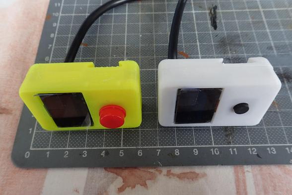 Zwei der neukonstruierten Displaygehäuse mit taktilem Taster. Die Gehäuse liegen seitlich auf einer Matte, Ansicht von schräg oben. Das Gehäuse der linken Displayeinheit ist in gelb gehalten mit einem roten Knopf (aufsetzbar / wechselbar). Die rechte Einheit wurde in weiß gedruckt, der Tastknopf ist schwarz und von der Größe her deutlich kleiner als der aufsetzbare Knopf links. Ebenso ragt der schwarze Knopf nicht so weit über das Displaygehäuse.
In Höhe des Knopfes ist auf der Hinterseite die Schwalbenschwanzführung ausgenommen, etwa hinter dem Display läuft das schwarze Kabel nach hinten weg.