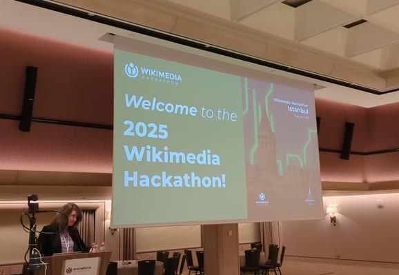 A presentation screen in a well lit beige hotel saloon displaying a green welcome to wikimedia hackaton 2025. A women presenting.