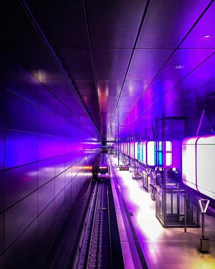 A modern subway station in Hamburg’s Hafencity, illuminated by striking purple and blue light panels. A train approaches the platform, surrounded by sleek architecture and glowing reflections.
