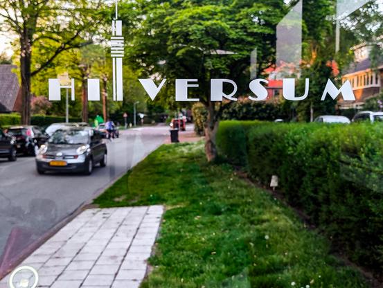 A photo taken from inside a bus stop, looking out onto a street. The word "HILVERSUM" is prominently displayed on the window. The text is a stylized white graphic that resembles the Hilversum Media Park tower.
