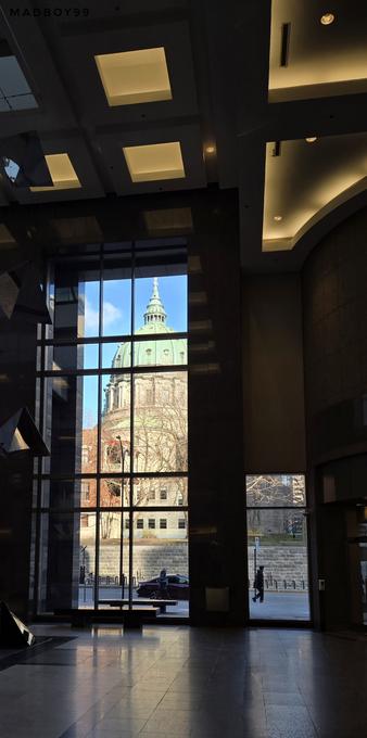 Picture of a basilica shot from the lobby of a bus station. Inside of the lobby is a bit dark, lights and forms can be seen on the ceiling, grey marble floor. At the left of the picture is a big window where the domed green roof (probably weathered copper) is majestically prominent, as well as part of the beige wall supporting it. Next to the right is a smaller window. Passerbys can be seen as well a part of a parked car.