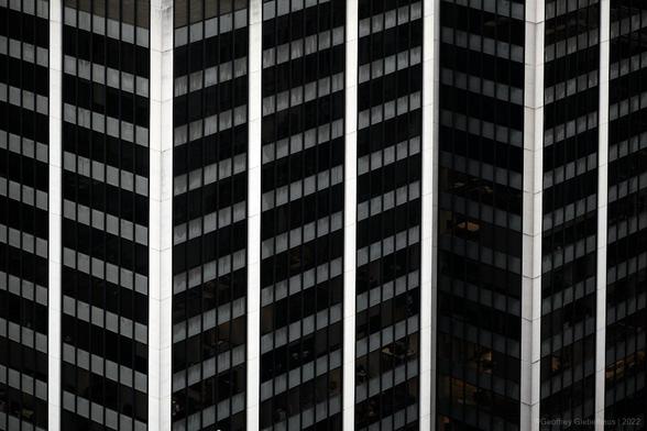 An almost abstract image looking at a series of buildings that have repeating white concrete columns. The layers of office towers creating a chevron styled zag pattern of black and grey lines across the image.
