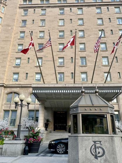 A front view of the bottom half of the Fort Garry Hotel in Winnipeg. The façade are bricks of many shades of beige and brown with many rows of windows, which are our hotel rooms. Below are stairs going up to the entrance to the hotel. The roof has 5 Canadian and American flagpoles jutting up and out, their flags blowing slightly in the wind. To the left of the stairs are a three-light lamp post and big pots of flowers. In front of the stairs, the nose of a black car peaks out from behind a Bellhut, an oval-shaped structure made of metal and glass with a cement base, used by valets and bellhops.