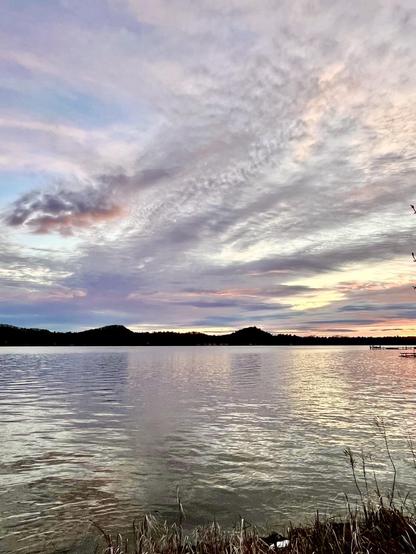 A serene lake scene at sunset, featuring calm waters reflecting colorful clouds and distant hills. The sky displays soft shades of blue, pink, and gray while grassy edges frame the view.