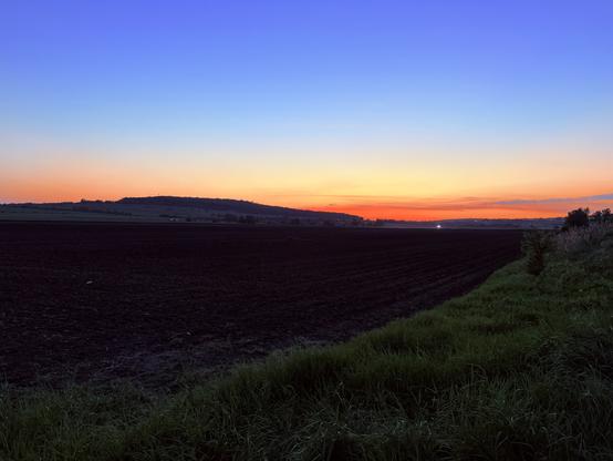 A serene landscape at dusk, featuring a gradient sky transitioning from blue to orange, with a silhouette of rolling hills in the background. The foreground showcases dark, plowed fields, bordered by lush green grass.