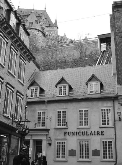 Black and white photo featuring a narrow street with historical buildings. In the foreground, a two-story building labeled "FUNICULAIRE" has multiple windows and decorative elements. Above, the funicular railway ascends a hill toward a prominent castle-like structure with towers and turrets. It is in fact the Chateau Frontenac hotel. A man and four kids can be seen in the foreground.