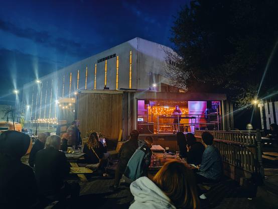 A pub garden aimed towards a stage inside a shipping container. A large industrial warehouse looms a little too closely to be background
