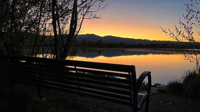 A metal park bench with small trees on either side overlooking a small, calm lake with barely a ripple in the water, the distant mountains across the lake a dark silhouette against the clear, post-sunset, dusk sky filled with a glowing gradient of golden-orange color, with the mountains and sky reflected in the glassy surface of the lake.