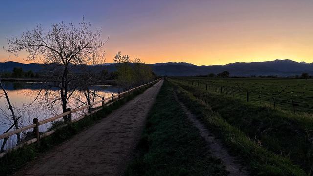 A dirt-gravel path alongside a small lake on the left and grassy fields on the right recedes into the far distance toward the silhouettes of far off mountains and the golden-orange glow of the clear, dusky sky.