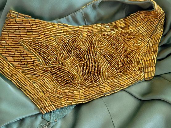 A shoulder of the dress laid out so you can see the detail of of the beadwork. A collection of 9 curved leaves in gold and brown seed beeds, overlaying each other. They are echoed in gold seed beads. Bugle beads swirl around them , in near orderly rows so they can catch the light.