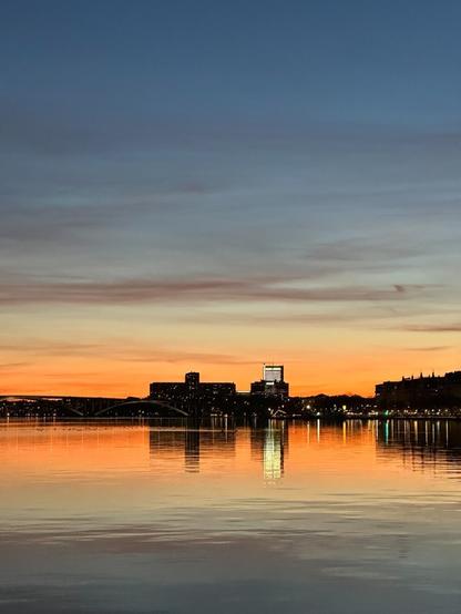 the sky, just after sunset, reflecting in a body of water. On a distant shore, there are buildings, including a brightly lit skyscraper. Thin wisps of cloud colour the sky like paintbrush strokes.