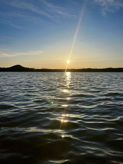 A serene sunset over a calm body of water, with gentle waves reflecting the golden light of the sun. Silhouetted hills are visible in the background against a clear sky.