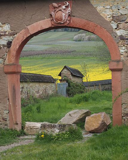 Farbiges Hochformat. Sogenannte intelligente Bots sehen ein Hufeisen. Menschen erkennen eine Bruchsteinmauer mit eingelassenem Torbogen aus Sandstein. Im Durchblick ist eine Wiese, eine weitere Mauer und ein Rebhang sichtbar.