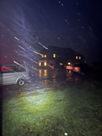 A parking lot with a couple of cars, a house and lots of rain in the night.