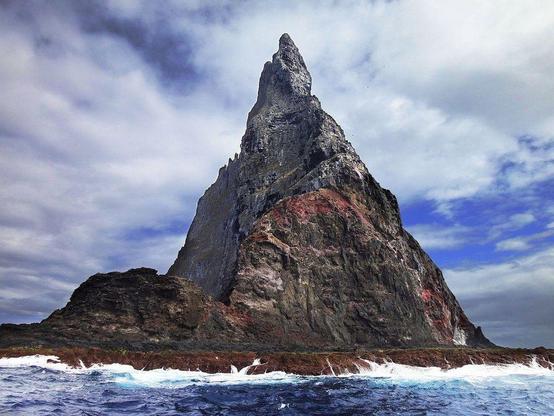 Vue majestueuse de la pyramide de Ball, un monolithe rocheux acéré s'élevant au-dessus des flots dans le parc marin de Lord Howe. Sa silhouette imposante contraste avec le ciel nuageux, tandis que les vagues viennent s’écraser contre sa base escarpée. Les nuances de gris et de rouge sur la surface rocheuse renforcent son aspect dramatique et sauvage.
