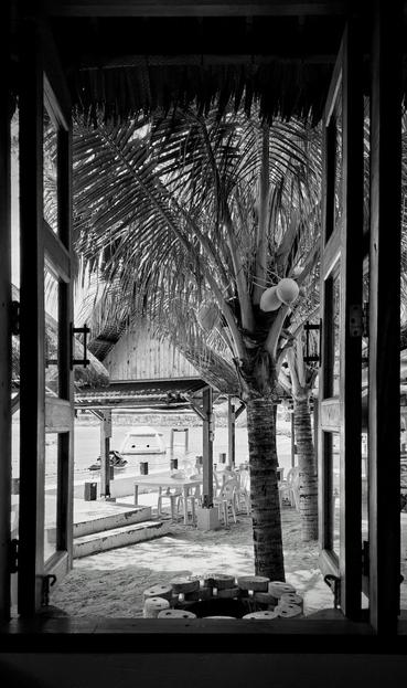 Black and white photo taken through an open window, framing a tropical beach scene with coconut trees, plastic chairs and tables on sand, nipa-roof structures, and a dock with a jetski and a blowup contraption floating on the water in the background.