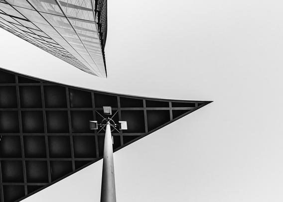Dramatic upward view of modern architecture featuring a tall glass skyscraper on the left and a triangular roof structure with a central streetlamp beneath it, all captured in stark black and white for a minimalist effect.