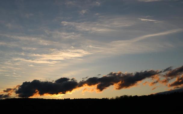 Photograph of a blue sky at sunrise over a dark horizon of trees. The sun is up and above the horizon but hidden behind a strip of low, grey and dense clouds, making them glow underneath with a golden light. Higher in the blue sky, horizontal white clouds stretch from side to side.