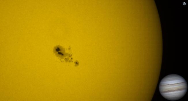 An archipelago of dark spots on the surface of the sun. They are surrounded by a slightly lighter region. Some mottled texture is visible on the sun; convection cells  the size of continents.

Off to the side are pasted images of Earth (which is unrecognisably tiny) and Jupiter (which is about 10 Earths across). The sunspot group is much larger than Jupiter.