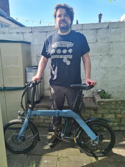 A man, facing the camera, standing behind a blue bicycle. His left hand is in the saddle, his right on the handlebars. 
The bike is a Fiido D11 and might look a bit small for him. (Such is the nature of folding bikes).
Behind him is a wall, a bit of a shed and a sliver of blue sky. He is partially bathed in sunlight. 
He is wearing a t-shirt with the words "Do not look to the dice to save you, they are not your friends," Woven between a selection of polyhedral dice, all showing the worst possible result.