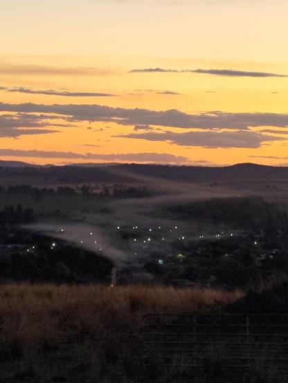 Sparse clouds in a flame-coloured evening sky above the twinkling lights of a small country town in a foggy valley with a grassy hill paddock in the foreground.