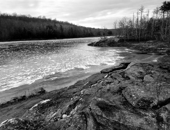 Monochrome image. On a bleak, cloudy and cold January evening, we are standing near the shore of a lake surrounded by a forest of leafless hardwoods with some pines and hemlocks. The lake is frozen solid, but there is not a flake of snow to be seen. Much of the near shoreline is fronted by large formations of jagged and serrated formations of dark gray rock speckled with light gray lichen and exposed veins of very light gray quartz.