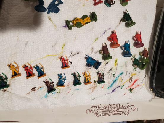 A photo of 8 tiny archers and 8 tiny spearmen laying on a paint-stained paper towel. Each of them has their clothes and some parts of their skin and armor painted in solid blocks of color. The spearmen are small compared to common minis, and the archers are even smaller.