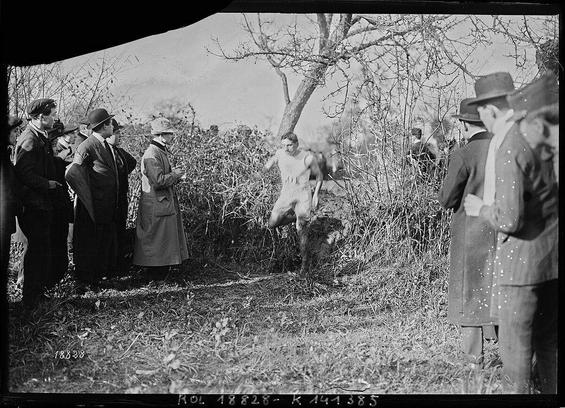 Photographie d'une course de cross-country en plein air. Jean Bouin, athlète vêtu d'une tenue de sport traverse un terrain herbeux, sous les regards attentifs des spectateurs alignés sur le bord du parcours. Certains observent de près, tandis que d'autres prennent des notes. Le paysage hivernal, avec ses arbres dénudés et son sol légèrement boueux, évoque l'intensité et l'effort physique du coureur.