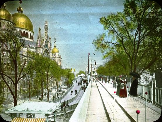 Photographie du trottoir roulant de l'Exposition Universelle de Paris 1900, une innovation technologique fascinante de l'époque. L'image montre des visiteurs se déplaçant sur une plateforme mobile entourée de balustrades blanches ornées de détails rouges. À gauche, des bâtiments majestueux aux dômes dorés, probablement le Palais des Beaux-Arts, dominent la scène. Des arbres verdoyants bordent le trottoir roulant, ajoutant une touche naturelle à cet espace urbain animé. Cette installation, surnommée la « Rue de l'Avenir », permettait aux visiteurs de parcourir l'exposition de manière fluide et rapide, incarnant l'esprit de modernité du début du XXe siècle.
