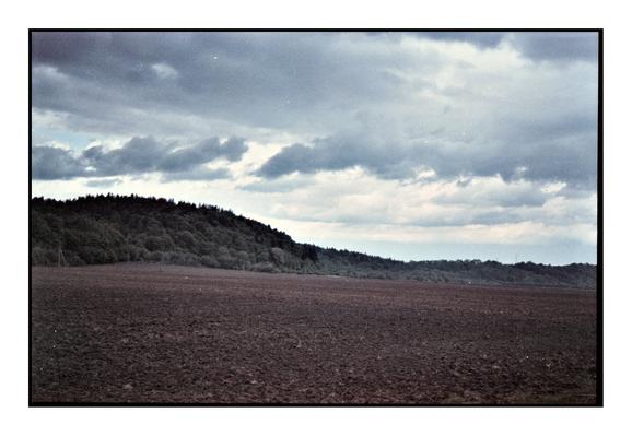 Grainy film photograph of a freshly plowed field under a dramatic, overcast sky with a forested hill in the background. Shot on expired Fujicolor 200 film with visible color shifts and speckles, emphasizing a moody, nostalgic aesthetic.