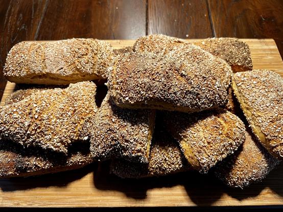 Auf einem dunklen Holztisch liegt ein Holzbrett. Darauf liegen zahlreiche frisch gebackene Brötchen mit rustikalem Ausbund