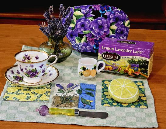 Tea tableau in green, yellow, and purple bringing together the flavors and visual appeal of lavender and lemons. On a green checkerboard tea towel are a purple floral cozy, purple flowered vintage china, a child's cup with painted lemons, a rock painted to look like a lemon slice, fabric painted to look like lavender sprigs, a small MidCentury Modern green glass vase holding dried lavender, and a box of Lavender Lemon herbal tea from Celestial Seasonings with an image of lavender fields and lemon groves in Provence.