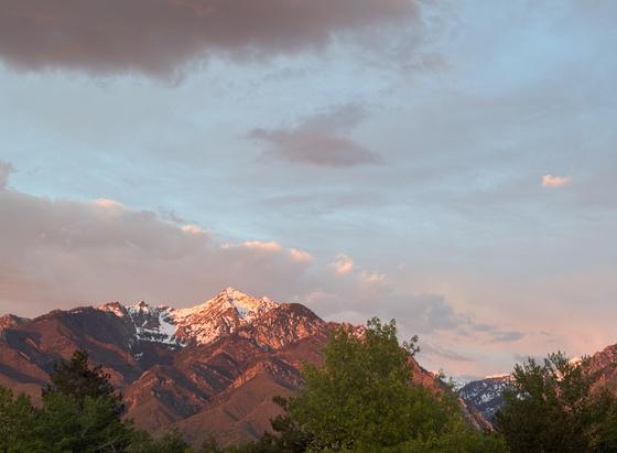 A view of the mountains seen from our balcony. The mountains are grey and brown granite, bathed in the red light of the sunset. They are tipped in snow. There are green trees in the foreground. The sky is a mix of shades of blue, grey, white, and pink.