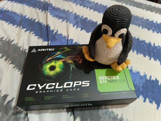 A photo of the box of an Artek mafe GeForce 750ti GPU on top of which there is an embroidered Tux plushie.