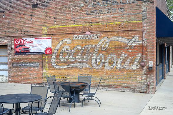 An old Coca-Cola ghost sign on a brick building next to a restaurant patio area.