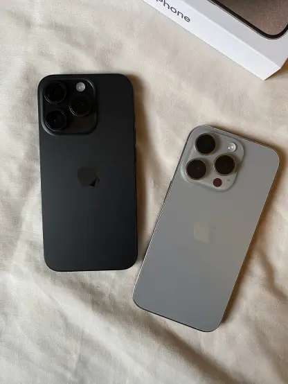 The back of 2 iPhone 15 Pro(s), one in Black Titanium and another in Natural Titanium - both in pristine condition, placed on a white bed sheet. In the top-right corner of the photo is a portion of the iPhone box.
