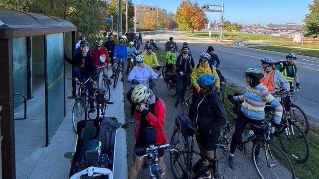 A group of people with, or on, their bikes is facing the viewer. They wear a variety of colourful clothes. The sky is clear on a sunny day.