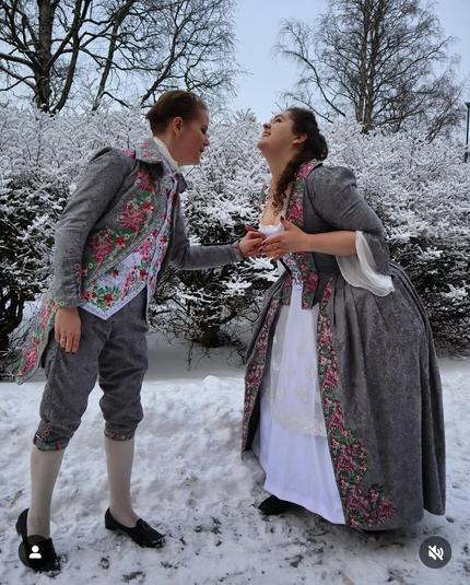 This image shows two people dressed in 18th-century-inspired historical costumes, standing in a snowy outdoor setting. The person on the left is wearing a gray and floral embroidered frock coat and knee-breeches with stockings and buckle shoes, resembling male aristocratic fashion of the time. The person on the right is in a matching gown with a similar floral motif, likely representing a woman's formal court dress from the same era.

Their joyful expressions and clasped hands suggest a playful or affectionate moment.