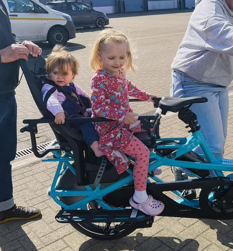 Two young girls aged 1 and 5, testing out the rear seating on a cargo bike. They are smiling and excited to be on the bike.