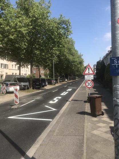 Eine Straße mit zwei Fahrspuren, getrennt durch weiße Streifen. Vor dem ersten weißen Strich steht eine rot-weiße Warnbarke. Rechts auf der Fahrbahn ist ein Bushalt markiert. Auf dem rechten Gehweg steht eine dreieckiges Achtung-Verkehrsschild mit Zusatzschild "Fahrbahnmarkierung fehlt" und einem Tempolimit von 30 km/h.