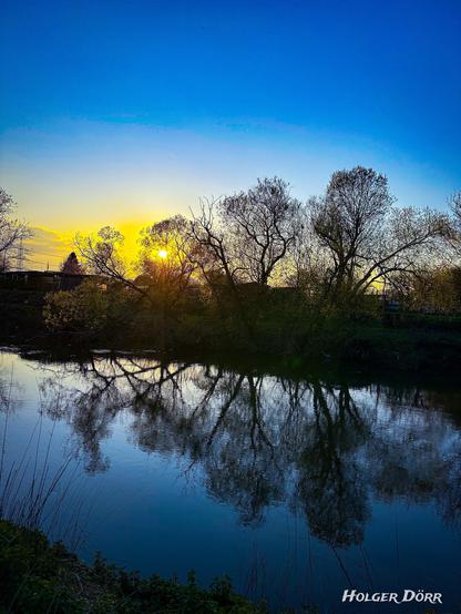 Ein ruhiger Frühlingsabend an der Lahn in Gießen. Die tief stehende Sonne wirft warmes, goldenes Licht durch die kahlen oder gerade austreibenden Äste hoher Bäume. Ihr Licht spiegelt sich sanft auf der glatten Wasseroberfläche des Flusses. Die Szene wirkt still, friedlich und leicht verträumt.