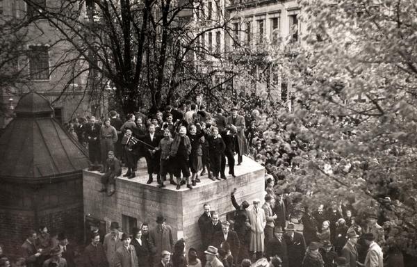 A black and white photo of a public square in Bergen, filled with people. In the middle, a small concrete building, a nazi bunker, with children with Norwegian flags on top, celebrating.